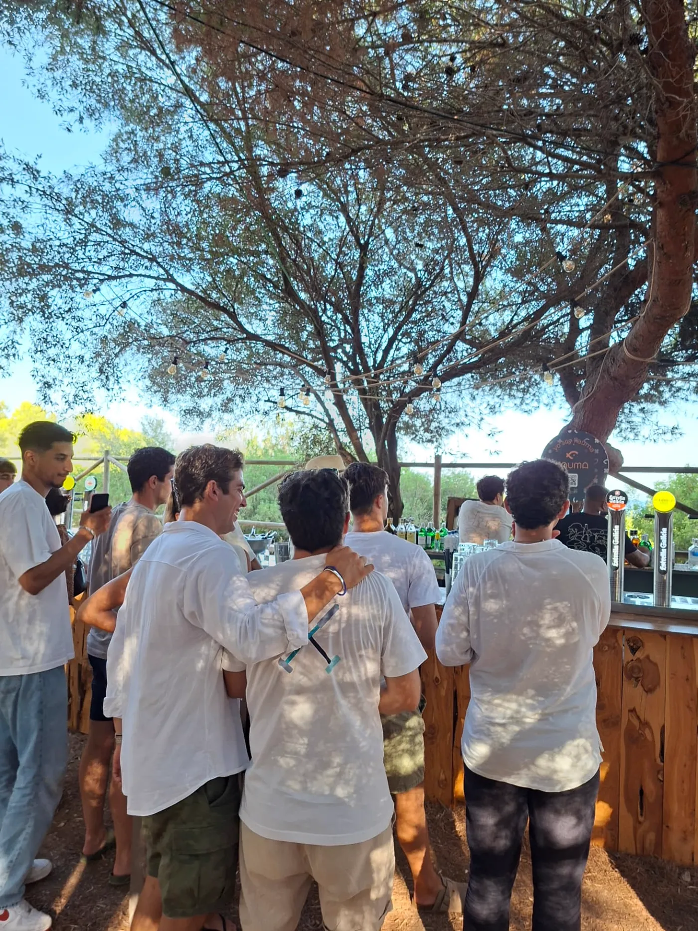 Grupo de personas vestidas de blanco charlando junto a la barra de madera de Bosque, bajo los pinos y las bombillas.